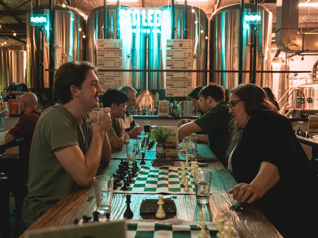 Players at chess boards along the wooden tables at Sea Legs Brewing Co, with the brewery's steel tanks and neon sign behind them.